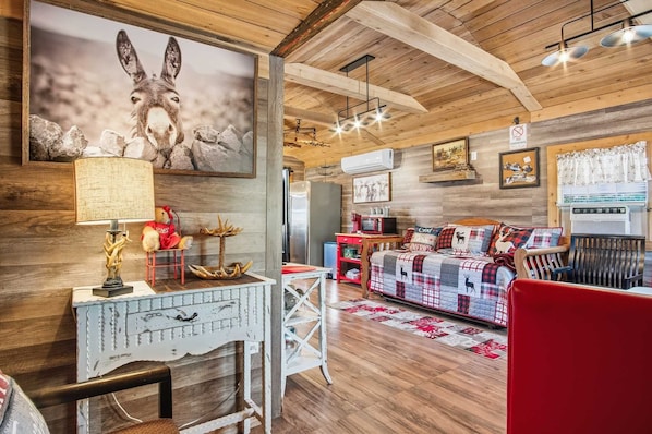 A cozy cabin interior with a wooden ceiling, a dining area with a table and chairs, a sofa with a plaid blanket, and a large painting of a deer.