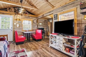 A cozy cabin interior with a wooden ceiling, a large TV, a dining table with red chairs, and a bookshelf filled with books and decorative items.