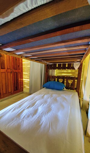 A single bed with a white sheet and a blue pillow in a room with wooden beams and a sloped ceiling.