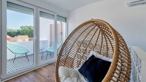 A room with a wicker chair, a bed, and a view through large sliding glass doors to an outdoor area with chairs and a tiled floor. - Minty House (Setúbal)