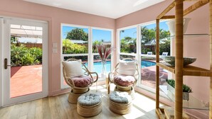 Quarto com cadeiras e pufes de vime, prateleira de vidro e vista para uma piscina.