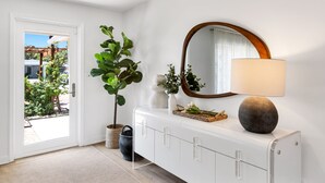 Uma porta branca que dá para um jardim, um vaso de planta, um espelho, um abajur e um armário branco com puxadores transparentes.