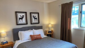 Cama bem arrumada com colcha xadrez, duas almofadas e uma almofada decorativa com texto. Duas silhuetas de animais emolduradas na parede, mesa de cabeceira com abajur e livros, e janela com cortinas.