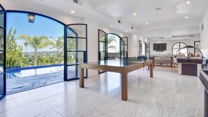 Ein geräumiges Zimmer mit einem großen Tisch, einer Couch und einem Fernseher. Große Fenster bieten Blick auf Palmen und einen Pool.