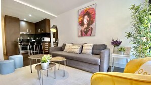 Ein modernes Wohnzimmer mit einem grauen Sofa, einem gelben Sessel und einem Couchtisch mit Pflanzen. Im Hintergrund ist eine Küchenzeile mit dunklen Schränken und ein Esstisch zu sehen.