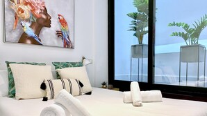 Ein ordentlich bezogenes Bett mit weißen Leinentüchern und aufgerollten Handtüchern, ein großes Fenster mit Blick auf Grünflächen und ein Gemälde einer Frau mit Papageien an der Wand.