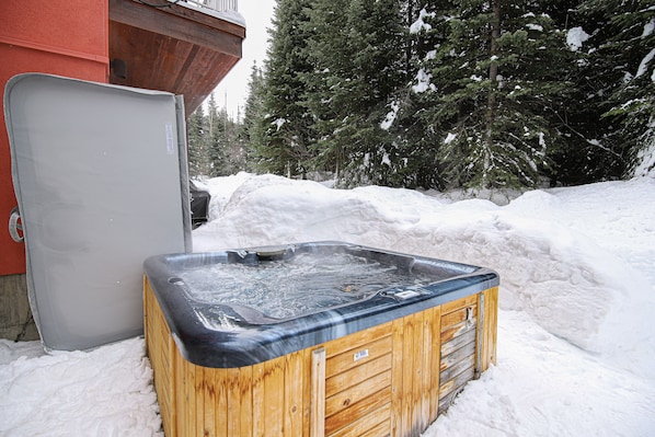 A wooden hot tub in a snowy outdoor setting with a red building in the background.