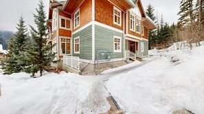 A two-story wooden house with a green and brown exterior, surrounded by snow and trees. - Settler's Crossing #13 | 3 Peaks Rentals (Sun Peaks)