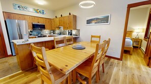 A modern kitchen with wooden cabinets, a stainless steel refrigerator, and a microwave. There is a dining area with a wooden table and chairs. - Settler's Crossing #13 | 3 Peaks Rentals (Sun Peaks)