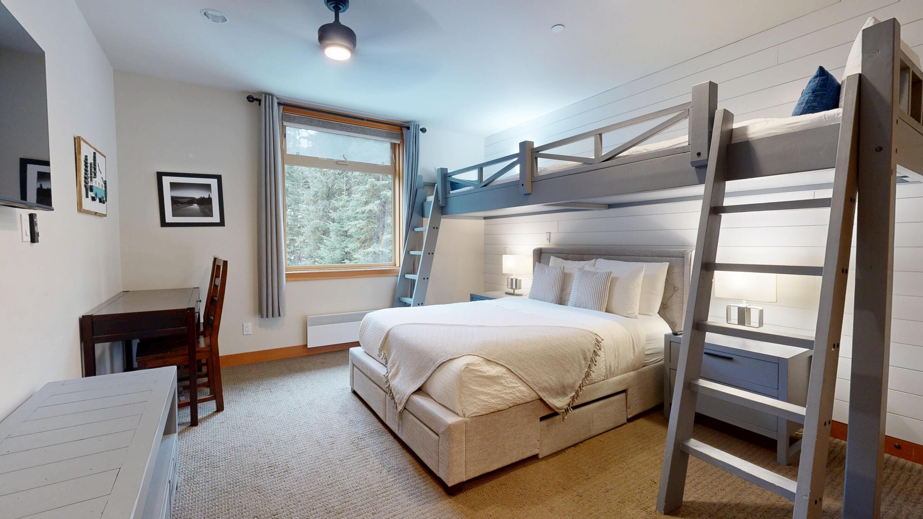 A bedroom with bunk beds, a desk, a chair, a window with a view of trees, and a ceiling fan.