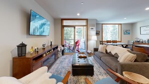 A spacious living room with a large TV, a fireplace, a sofa, and a dining area with a table and chairs. - Spacious Ski in/out Condo in Sun Peaks Village (Sun Peaks)