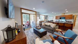 A modern living room with a sofa, ottoman, and chairs, a kitchen with wooden cabinets, and a sliding glass door leading to a deck. - Spacious Ski in/out Condo in Sun Peaks Village (Sun Peaks)