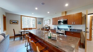 A modern kitchen with a central island, wooden cabinets, stainless steel appliances, and a large window offering a view of trees. - Spacious Ski in/out Condo in Sun Peaks Village (Sun Peaks)