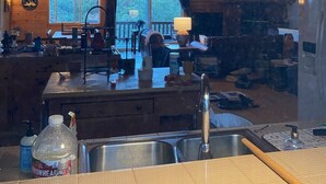 A rustic cabin interior with a wooden ceiling, a large kitchen island, a dining area, and a view of the outdoors through a window.