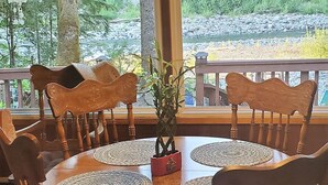 Une table de salle à manger ronde en bois, surmontée d’un set de table en fibres tressées, est entourée de chaises en bois. Un vase rouge contenant une plante est placé au centre.