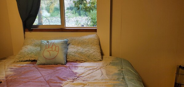 Una cama con un cojín mullido y una colcha, una ventana con vistas a vegetación, y un cabecero de madera.