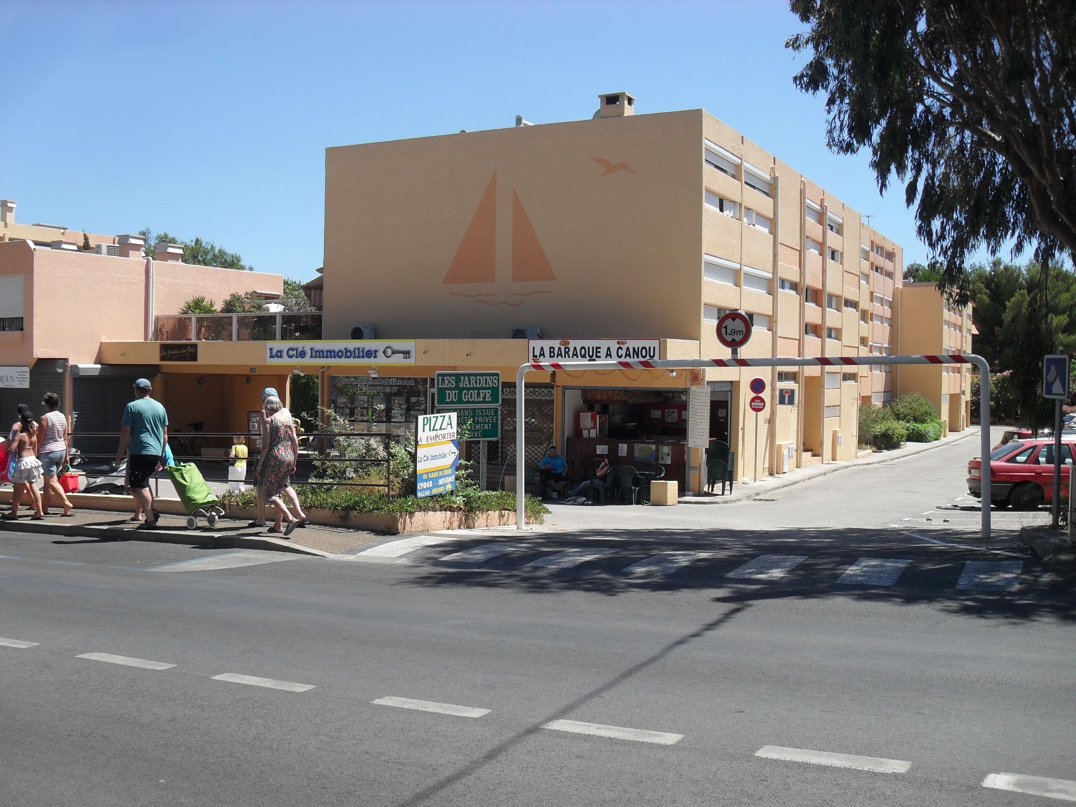 Vista de la calle con un edificio que muestra el logo de un velero, un letrero de pizzería y un letrero de restricción de altura de 1.9 metros.