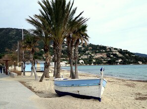 A beach with a row of palm trees, a small boat, and a hillside with buildings in the background.