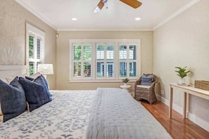 A bedroom with a large bed, a window with multiple panes, a ceiling fan, and a small side table.