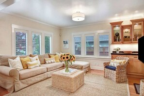 A living room with a beige sofa, a wicker coffee table, and a wicker chair. There are windows with blinds, a ceiling light, and wooden cabinets.