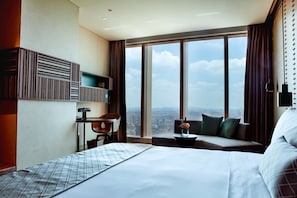 A hotel room with a large bed, a desk with a chair, a sofa, and a view of the city through large windows.