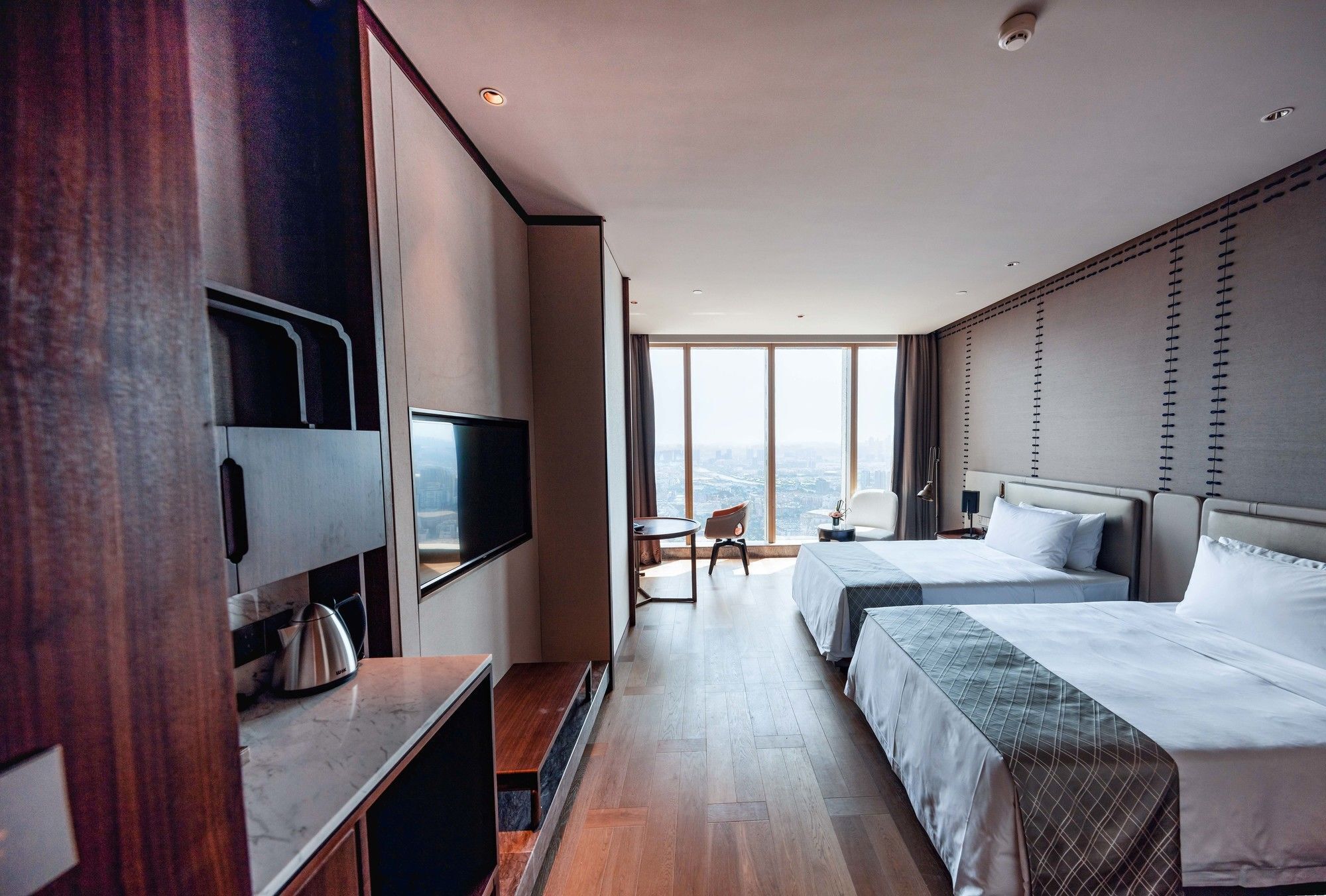 A modern hotel room with two beds, a television, a small table, and a view of the city.