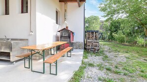 Ein rustikaler Essbereich im Freien mit Holztisch und -bänken, einem Steinofen und einem Garten mit Bäumen und einem Kiesweg.