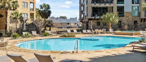 Une piscine avec des chaises longues, des palmiers et un immeuble à plusieurs étages doté de balcons.