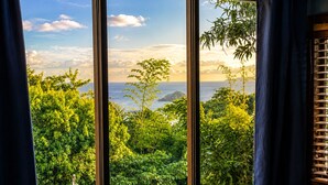 A room with a large window offering a view of lush greenery and a distant island. - Breathtaking Ocean Vistas, Jungle Bliss, Walk to Eateries & Beach, Fast Internet (Manuel Antonio)