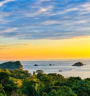 A coastal landscape with a sunset, rocky outcrops, and dense greenery.