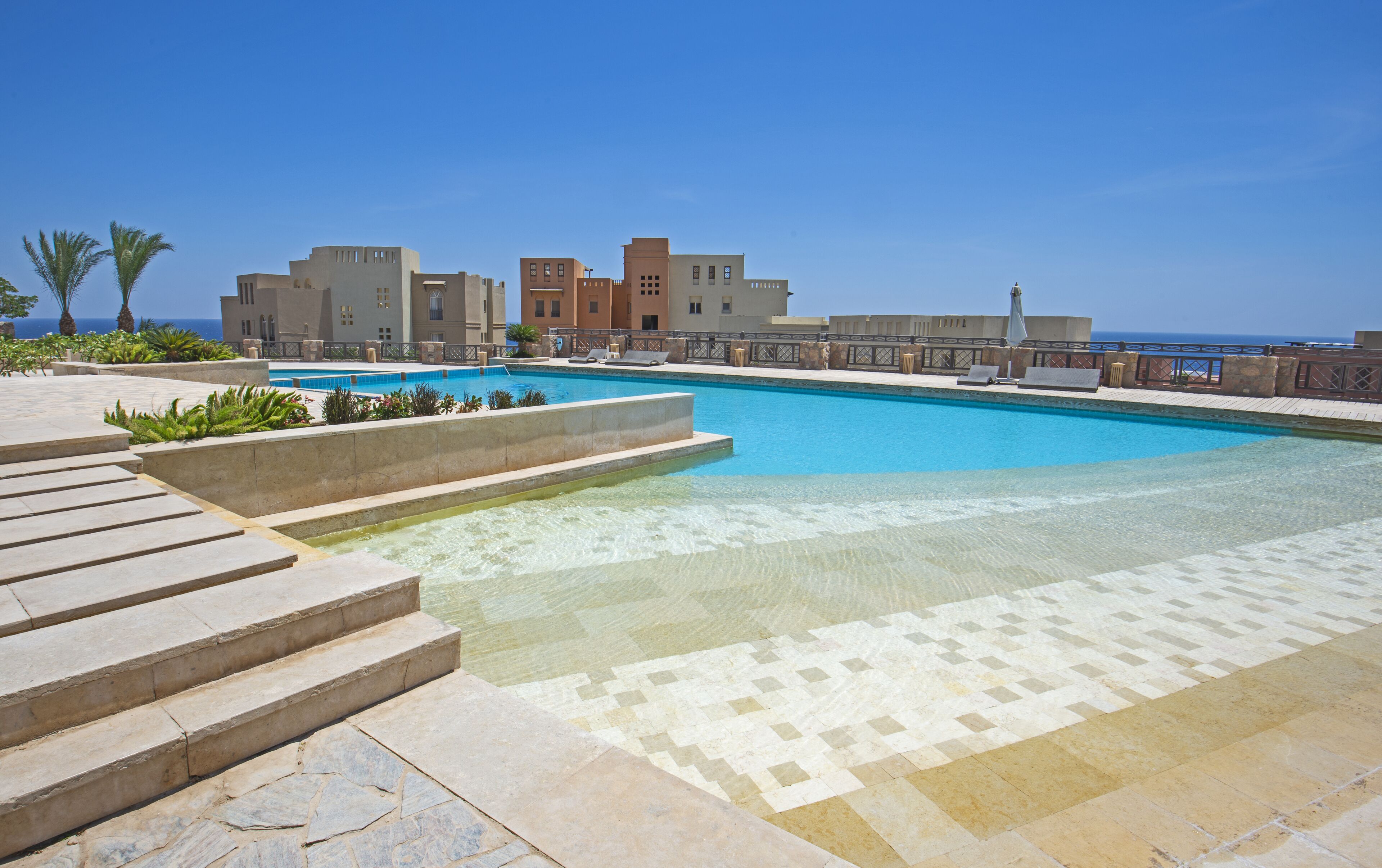 Ein Schwimmbecken mit klarem, blauen Wasser, umgeben von einer Steindeck und Stufen, die zum Wasser führen. Im Hintergrund sind Gebäude im modernen architektonischen Stil zu sehen.