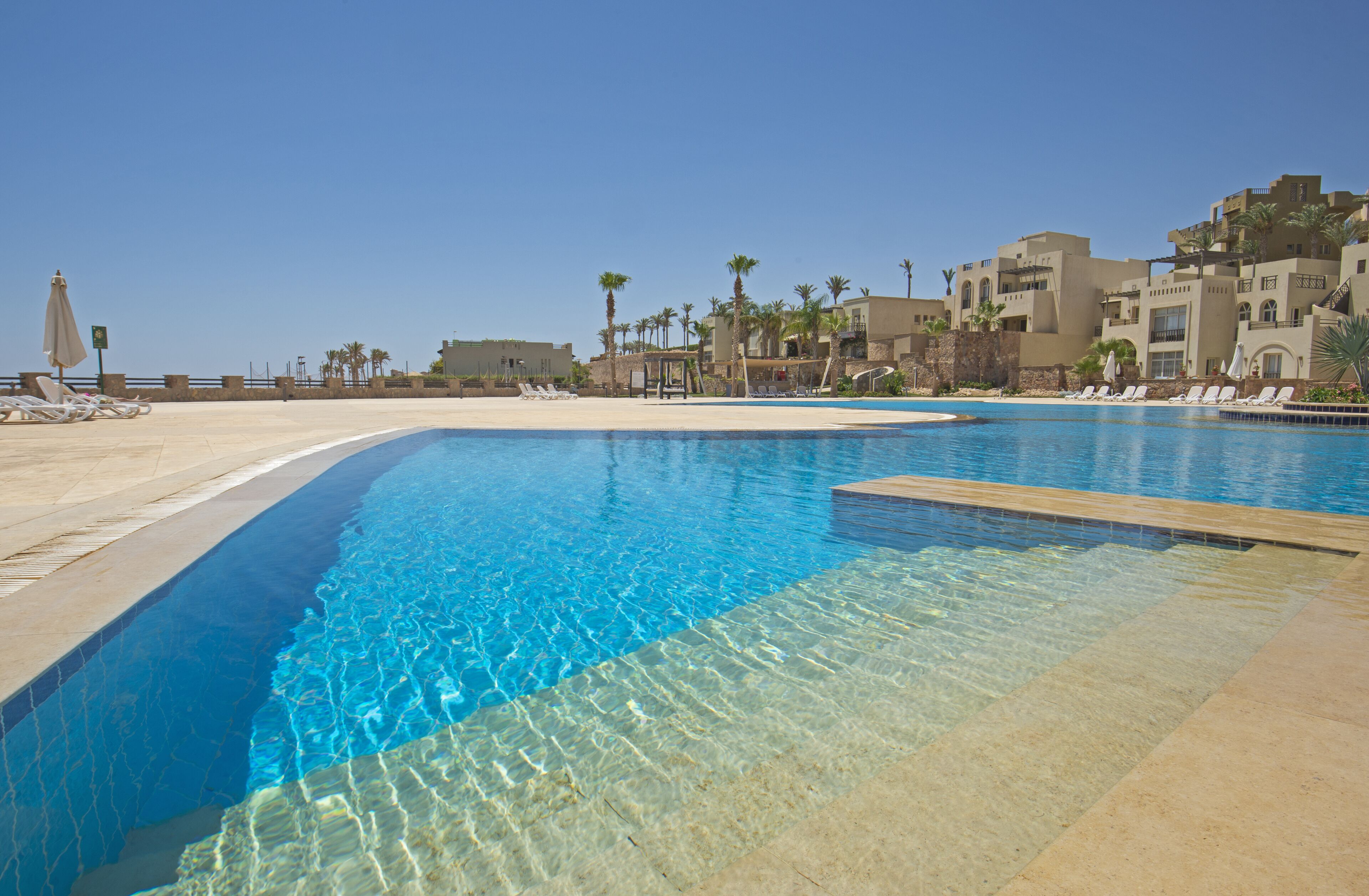 A swimming pool with clear blue water, surrounded by lounge chairs and palm trees, with buildings in the background.