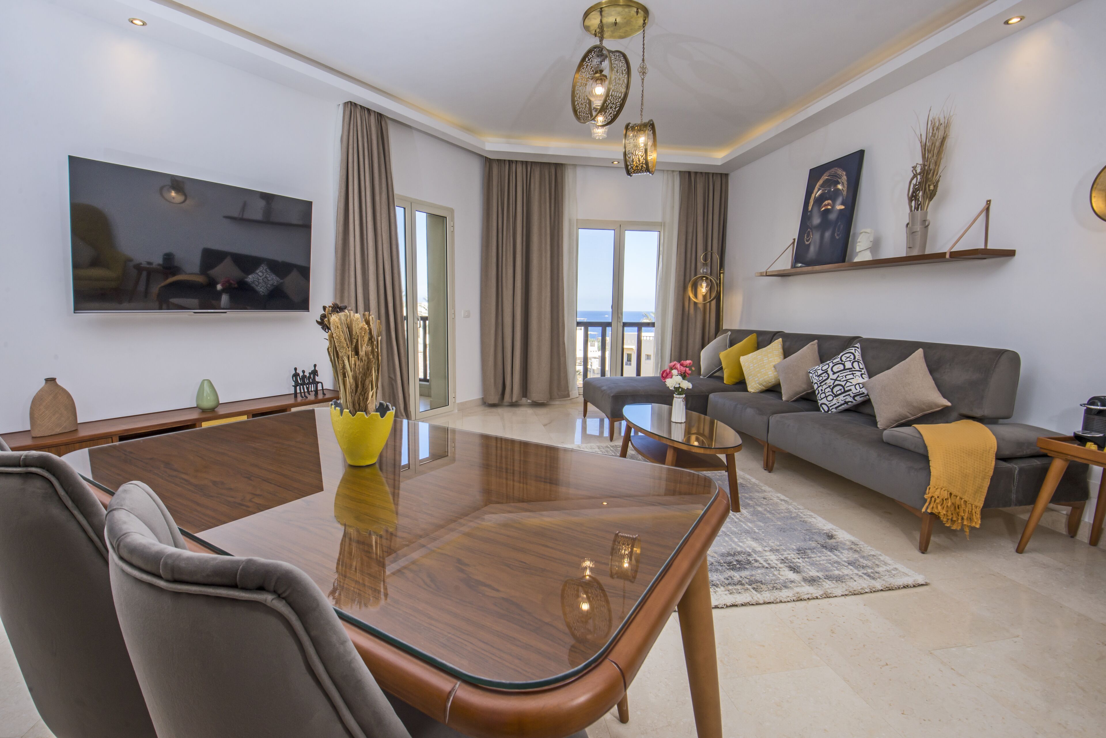A modern living room with a glass-top dining table, grey sofa, and a large flat-screen TV mounted on the wall.