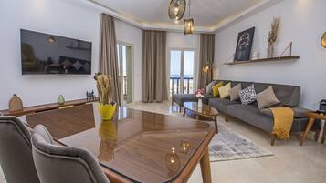 A modern living room with a glass-top dining table, grey sofa, and a large flat-screen TV mounted on the wall.