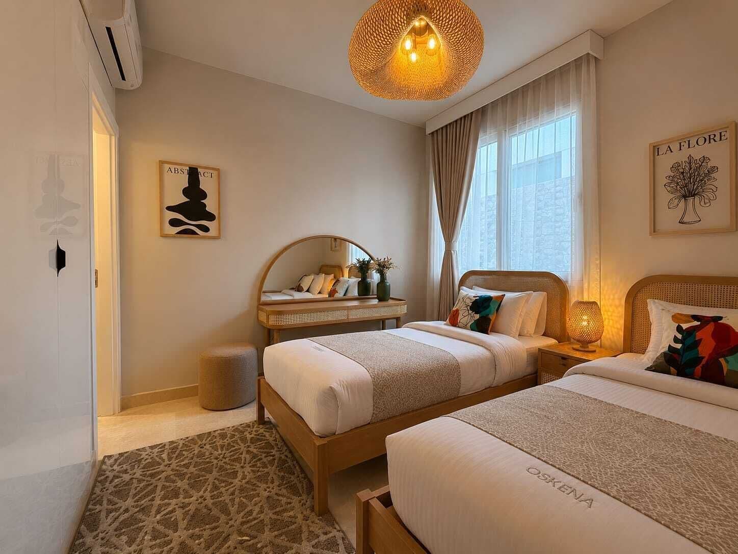 A hotel room with two beds, a bedside table with a lamp, a mirror, a wall-mounted artwork, and a window with curtains.