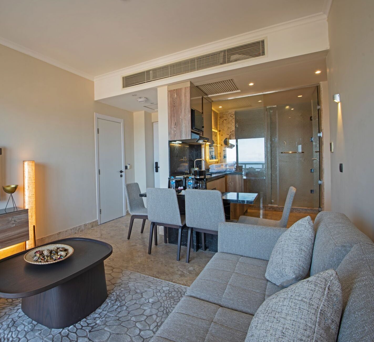Una sala de estar moderna con un sofá, sillas, una mesa de centro y una cocina visible a través de una puerta de vidrio.