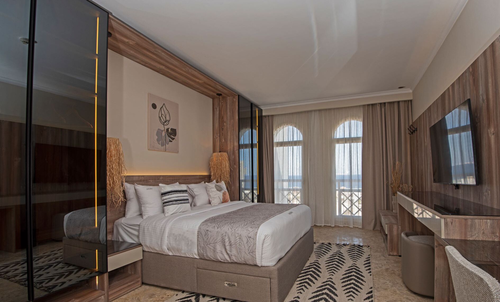 Ein modernes Hotelzimmer mit einem großen Bett, einem Fernseher, einem Schreibtisch und einem Balkon mit Vorhängen.