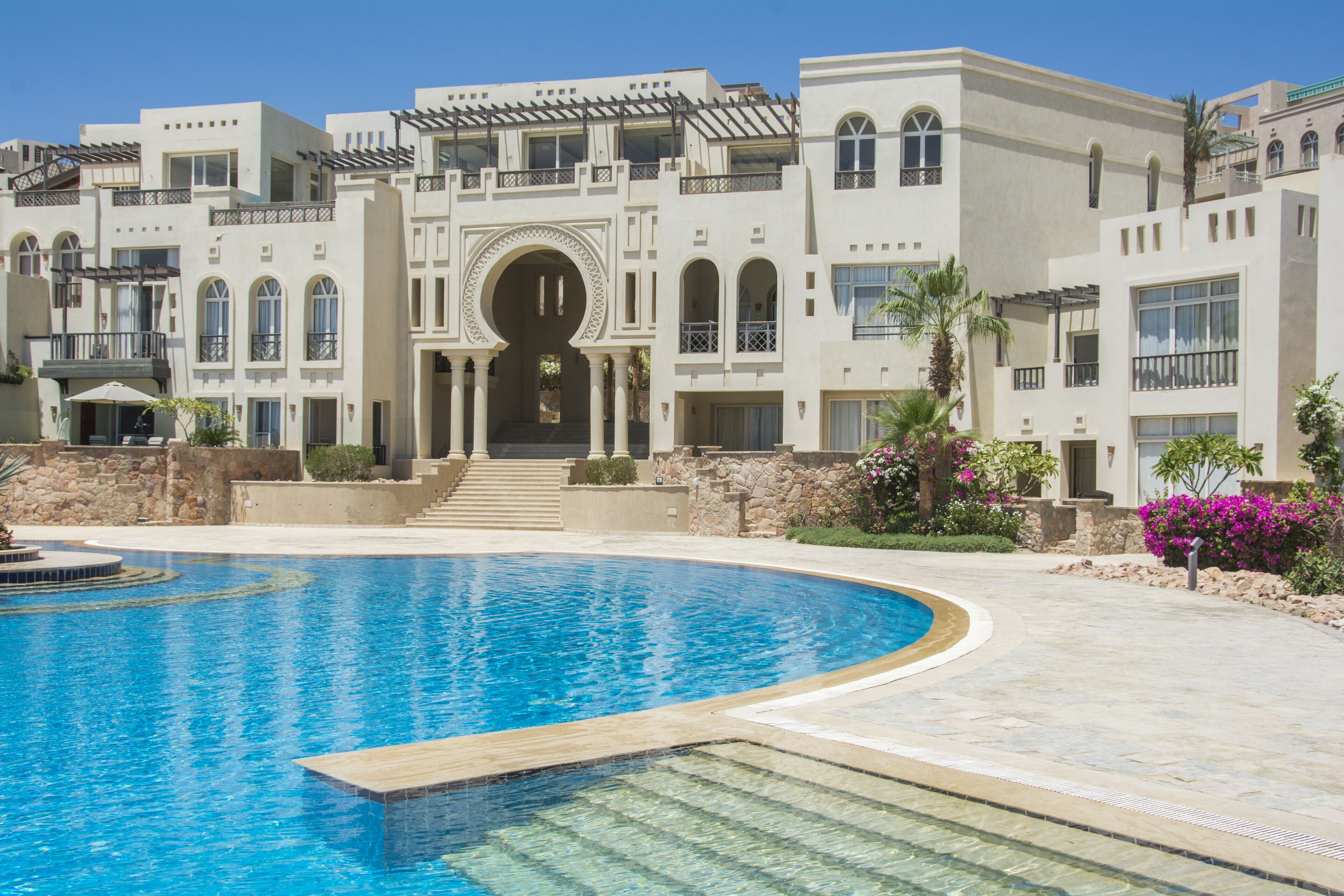 Una gran piscina con solárium de piedra, rodeada por un edificio de varios pisos con ventanas arqueadas y balcones.