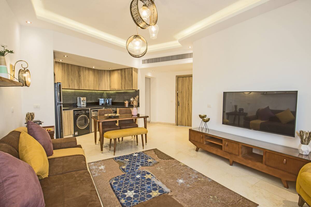A modern living room with a sofa, coffee table, and TV stand. There is a kitchen area with a dining table and chairs.