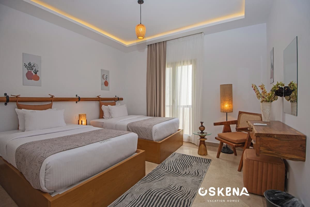 A modern hotel room with two single beds, a wooden desk, a chair, a small table, and a zebra-patterned rug.