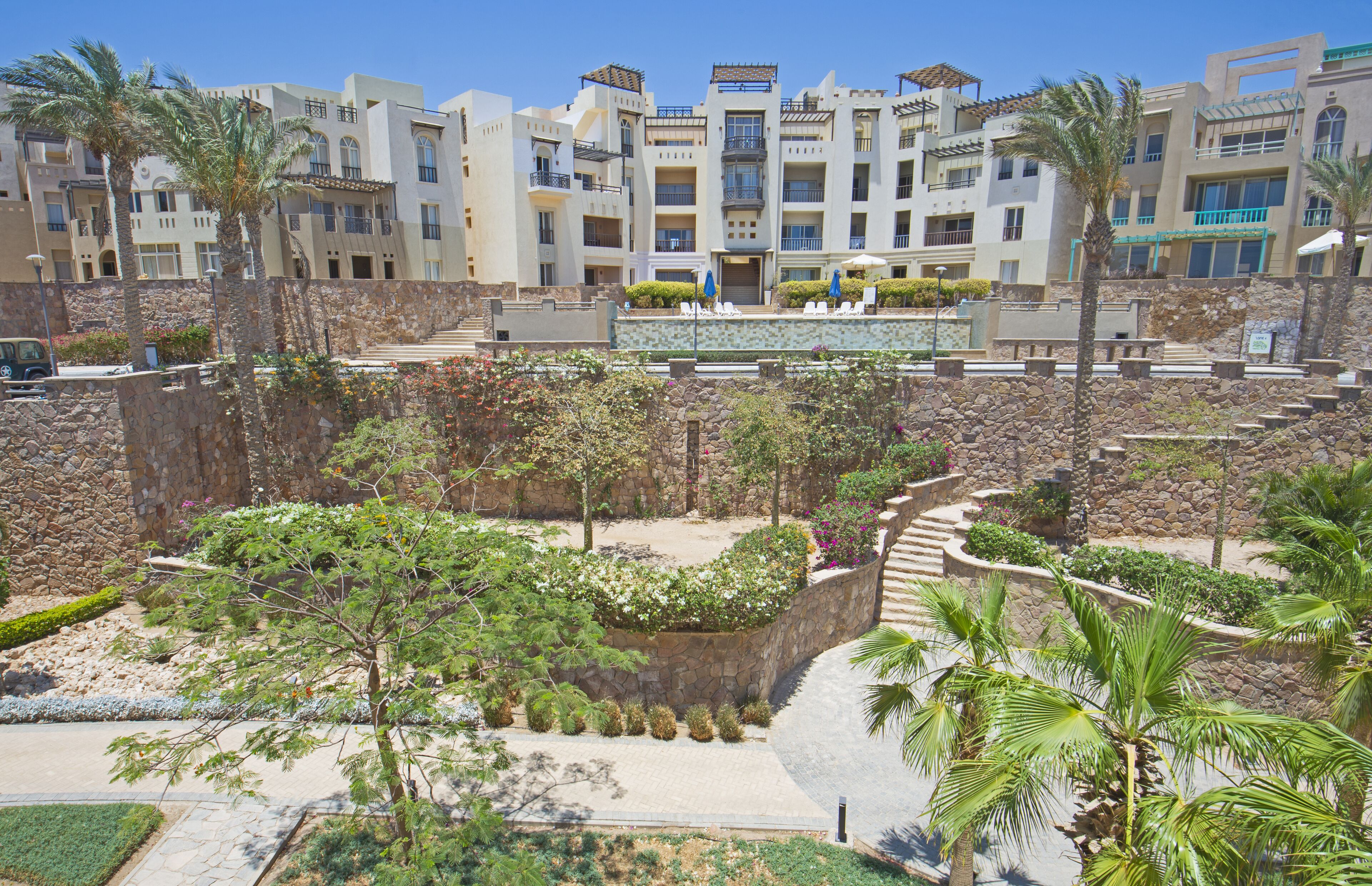 Un hotel de varios pisos con un patio ajardinado que presenta palmeras, un muro de piedra y un sendero que conduce a la entrada.