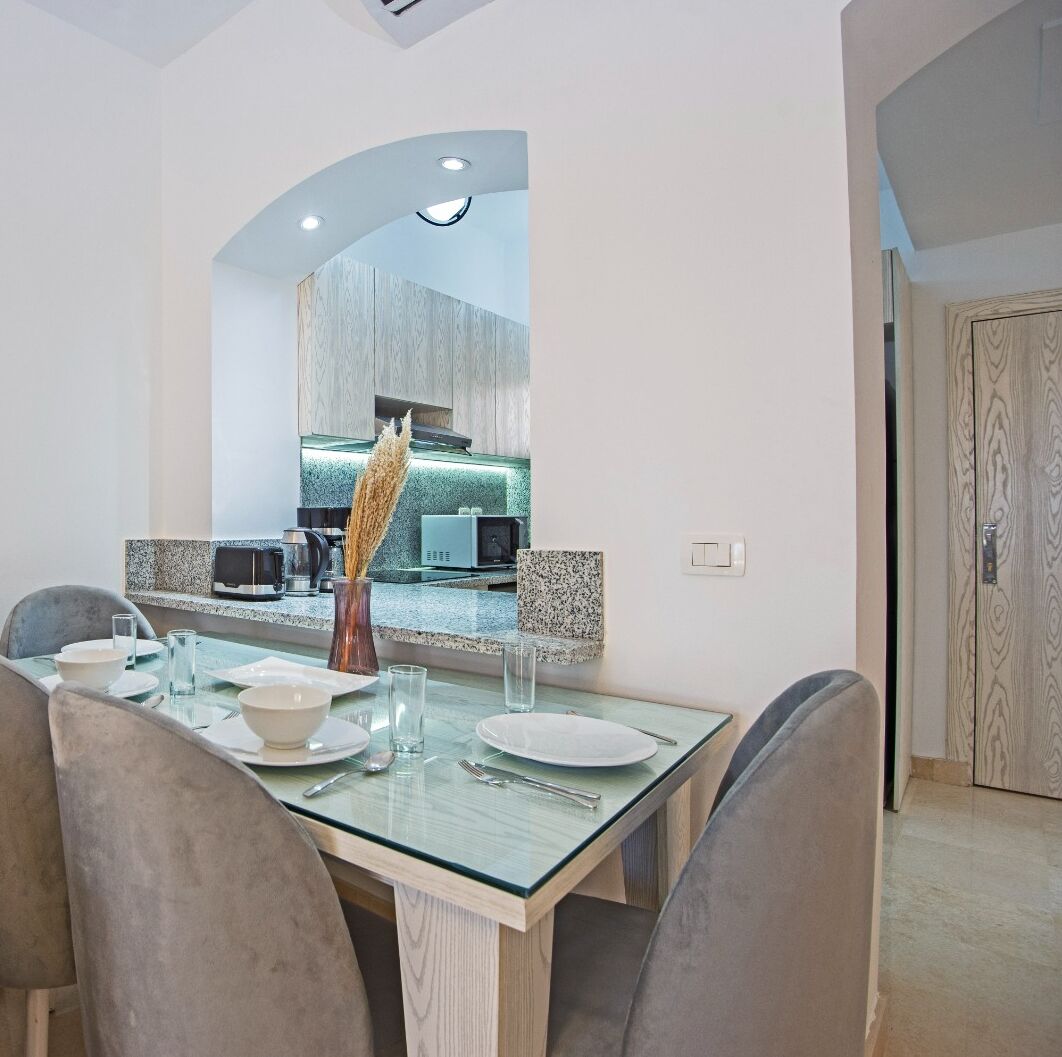 Un comedor con mesa de vidrio, sillas tapizadas de color gris y una cocina moderna visible a través de un arco.