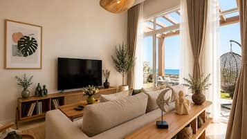 A modern living room with a large flat-screen TV, a sofa, a wooden coffee table, and a view of the ocean through large windows.