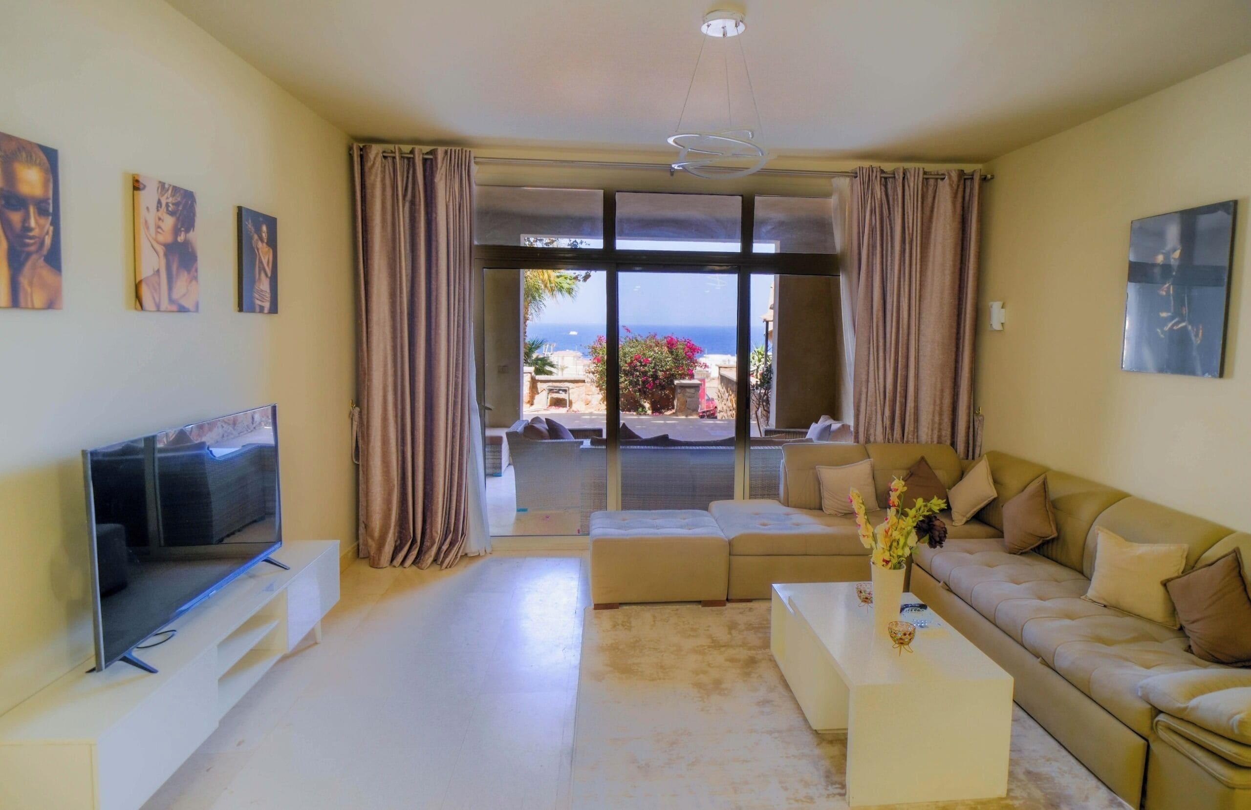 A modern living room with a flat-screen TV, a sofa, a coffee table, and a view of the sea through large windows.