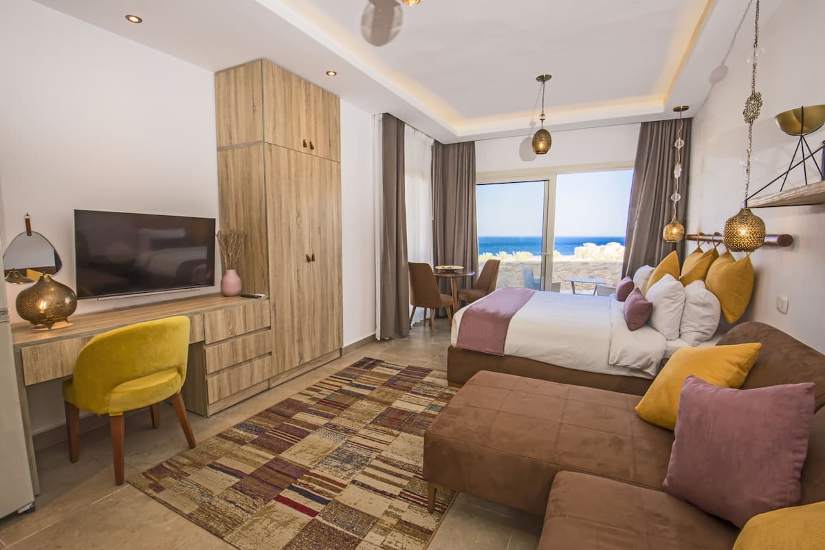 A hotel room with a large bed, a desk with a TV, a yellow chair, a patterned rug, and a view of the beach.