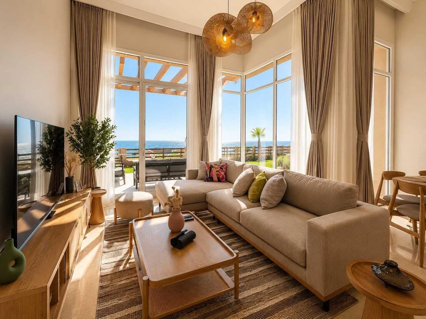 A modern living room with a sofa, coffee table, and TV, large windows with a view of the sea, and a balcony with outdoor furniture.