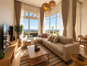 A modern living room with a sofa, coffee table, and TV, large windows with a view of the sea, and a balcony with outdoor furniture. - Outstanding Sea View-Azzurra Apartments (Sahl Hasheeh)