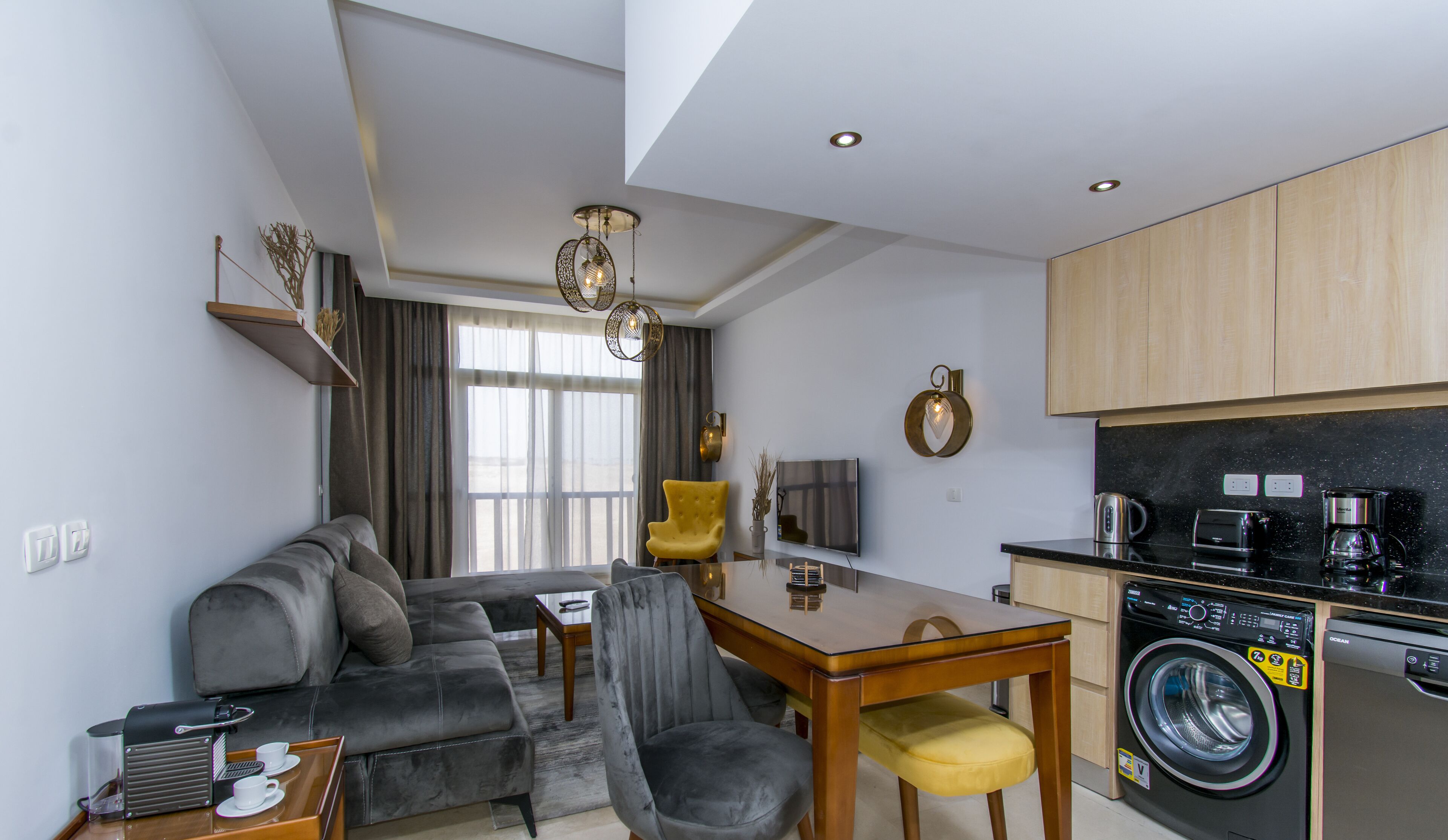 A modern living room with a sofa, a dining table, and a coffee machine. There is a washing machine in the corner.