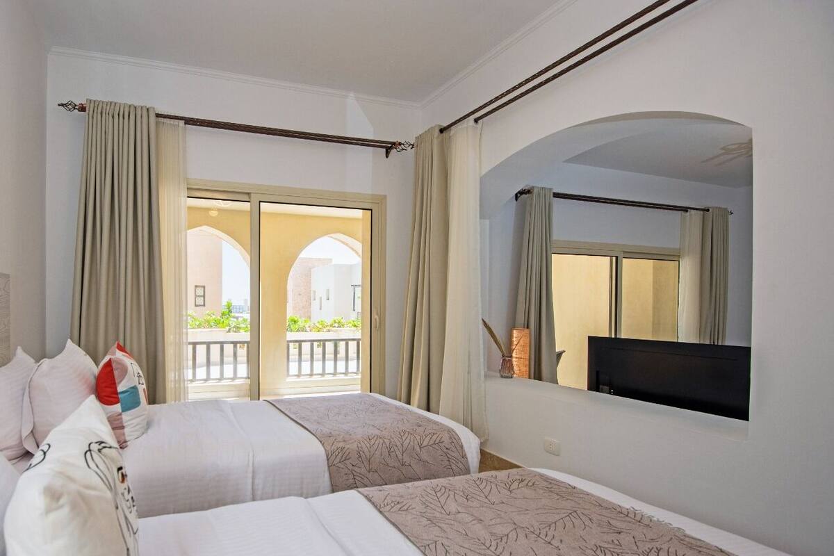 A hotel room with two beds, a large window with curtains, a flat-screen TV, and a balcony with a view of greenery.