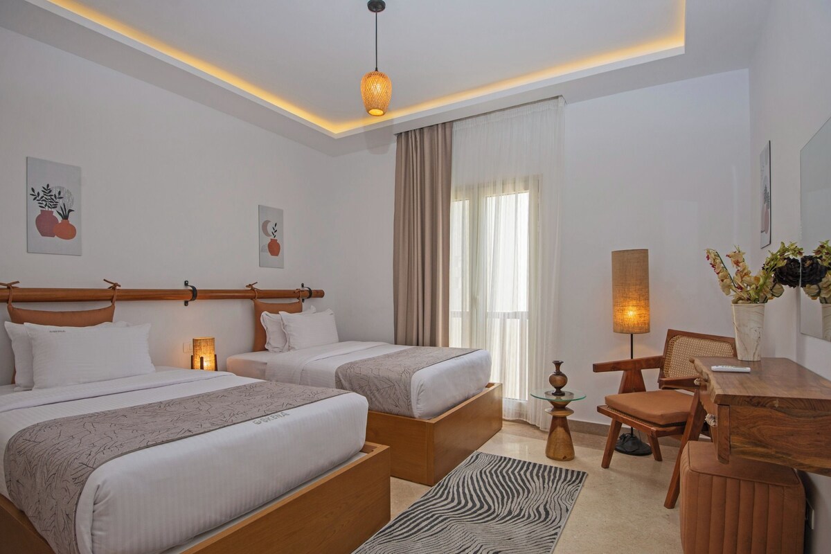 A modern hotel room with two single beds, a wooden desk, a chair, a small table, and a zebra-patterned rug.